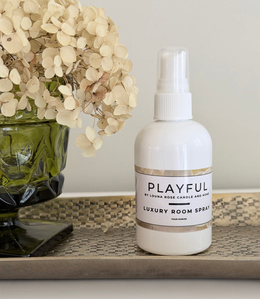 White room spray bottle labeled 'Playful' on a decorative surface with flowers.